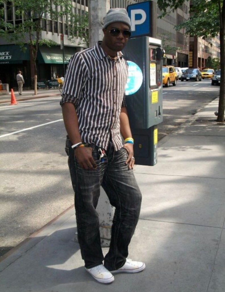 Young Black man standing on a New York City sidewalk in 2008, wearing a striped shirt and beanie, looking ahead with confidence and curiosity.