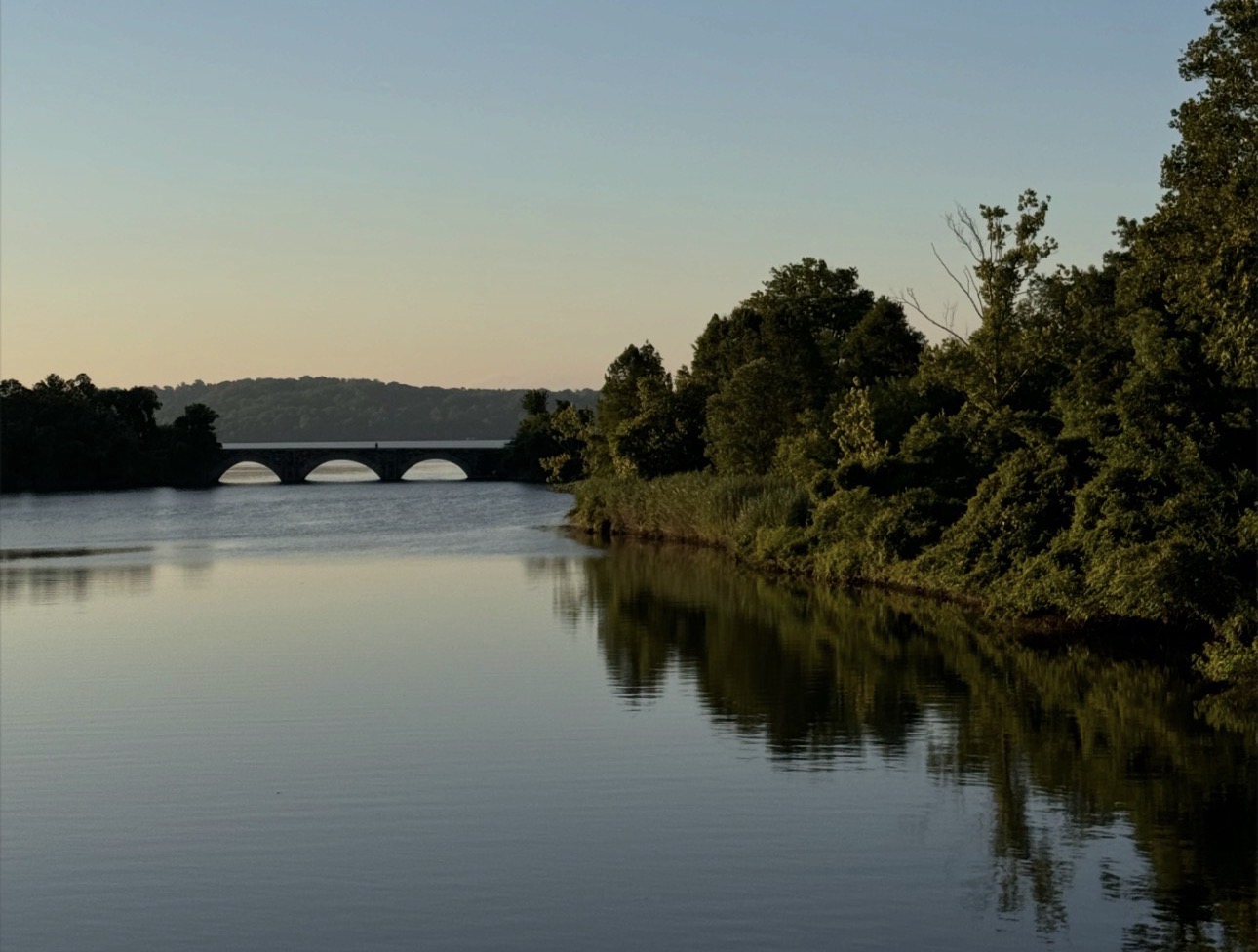 Calm river at sunset with trees reflecting in still water and a stone bridge in the distance
